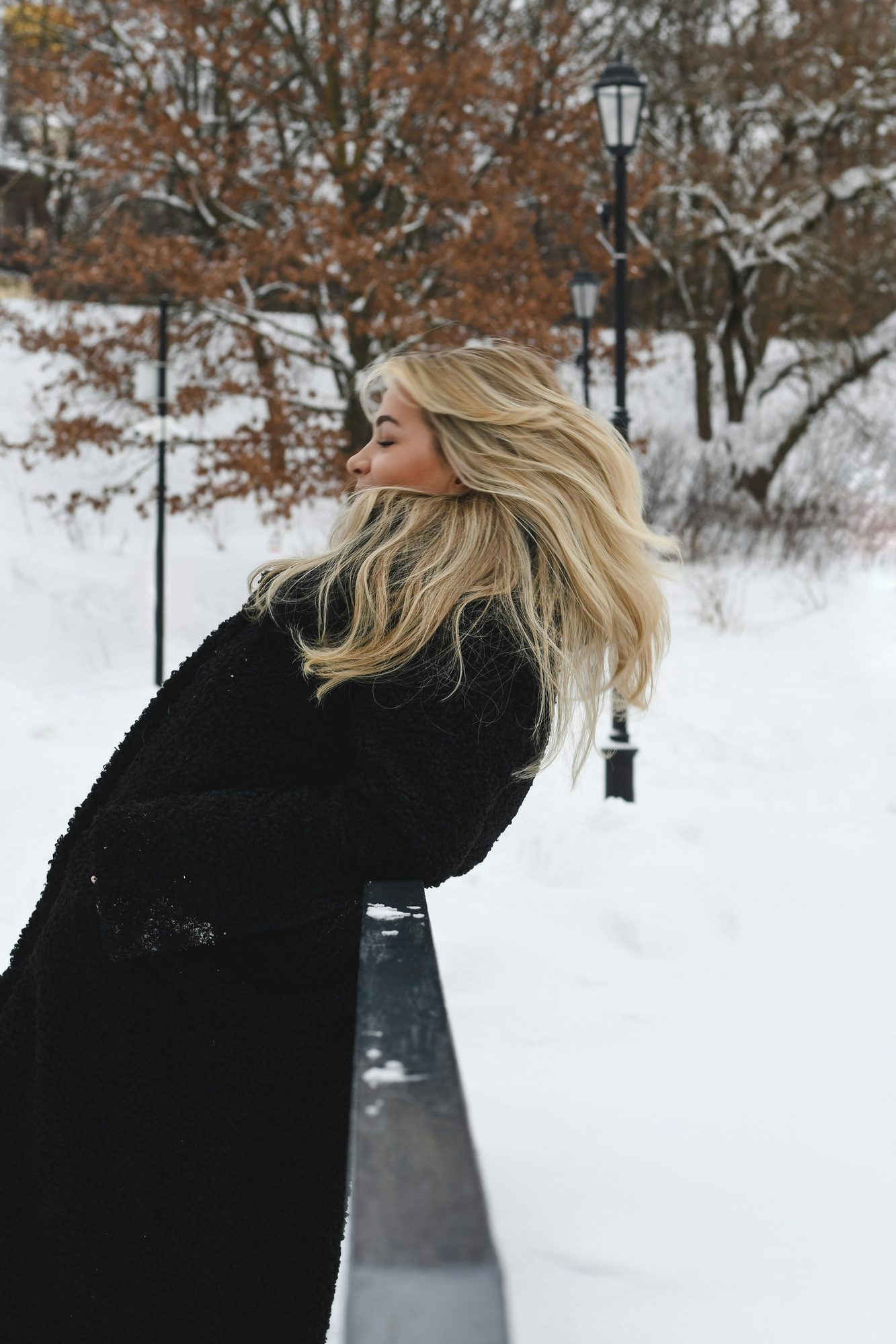 Mujer rubia con abrigo negro inclinada sobre una baranda en un parque nevado