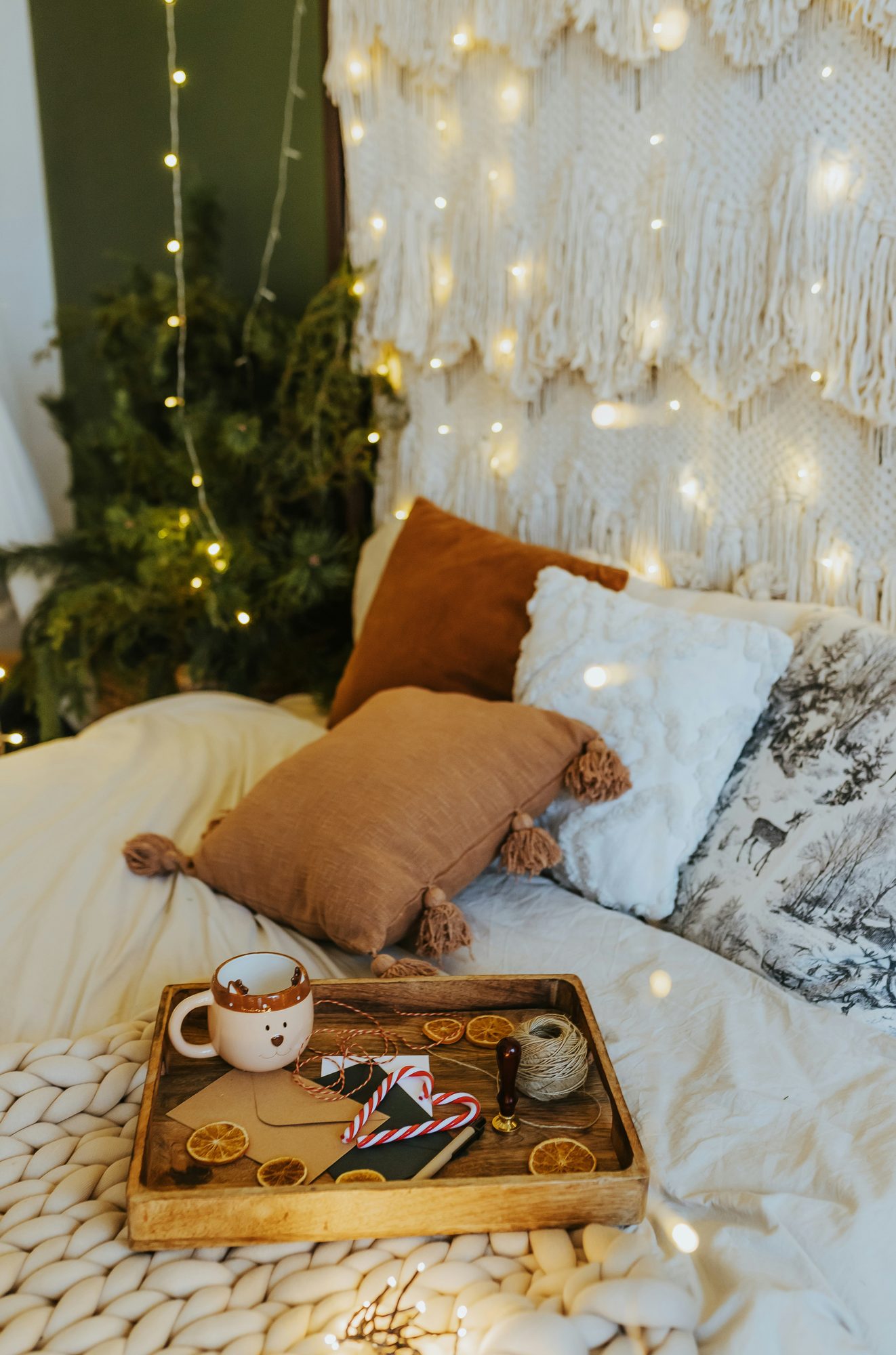Cama con cojines, manta de punto grueso y bandeja con taza bajo luces cálidas