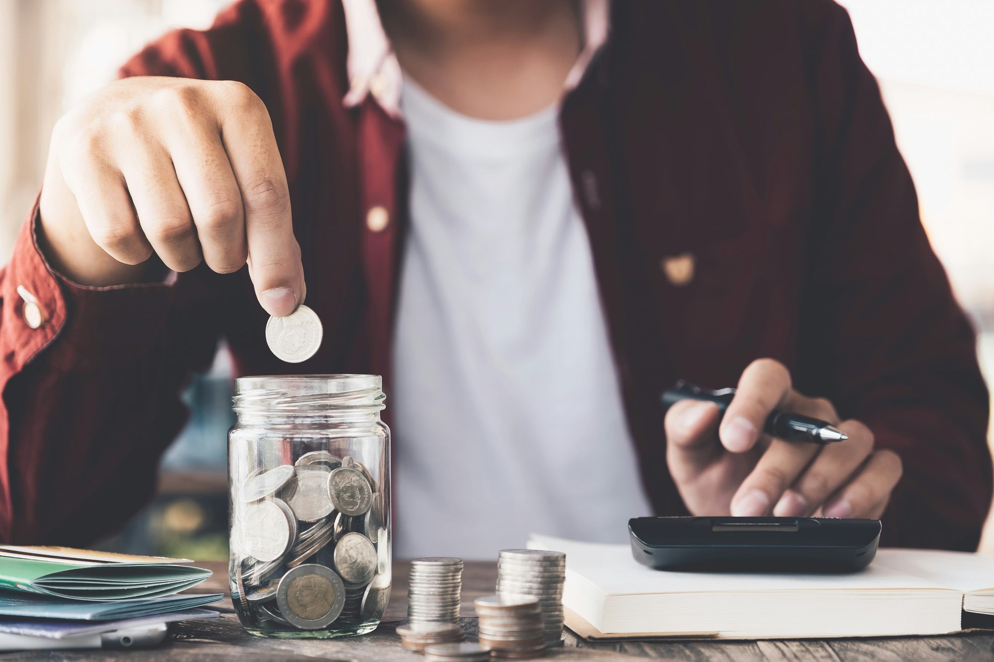 Manos poniendo monedas en un frasco de ahorro junto a una calculadora