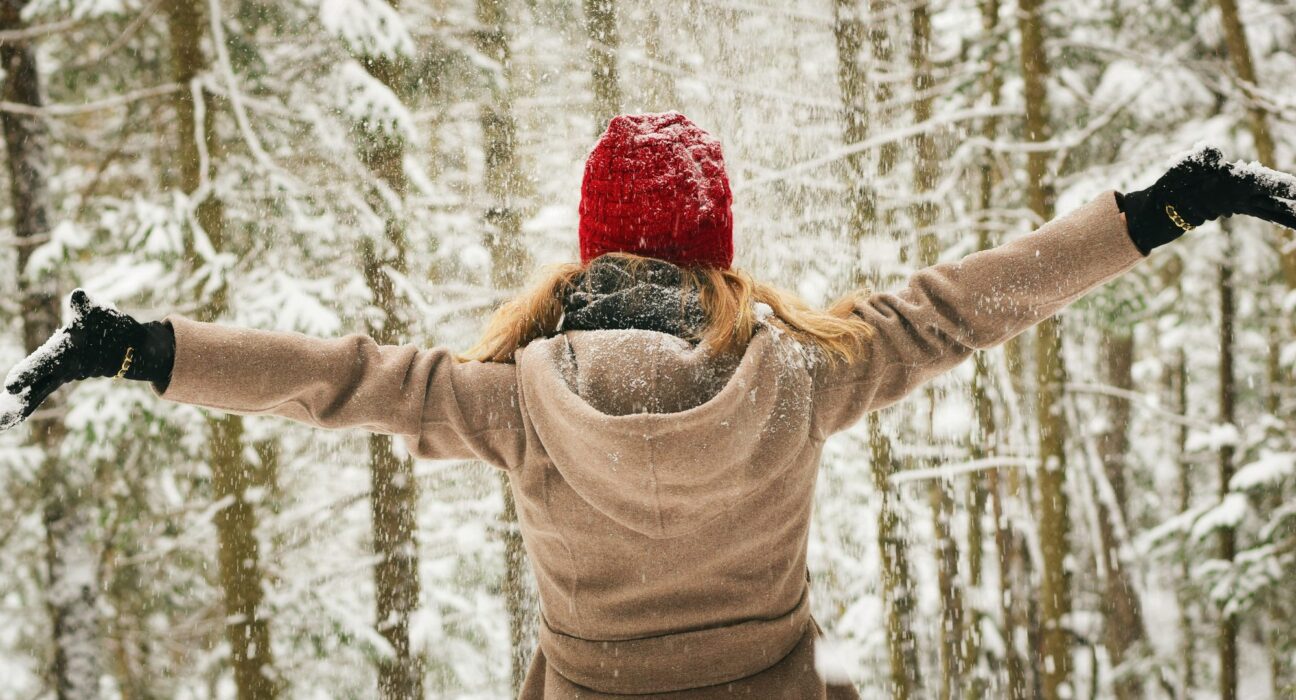 Mujer con gorro rojo y abrigo de espaldas, con los brazos abiertos en un bosque nevado