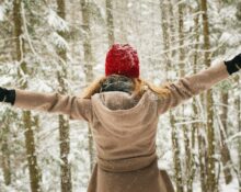 Mujer con gorro rojo y abrigo de espaldas, con los brazos abiertos en un bosque nevado