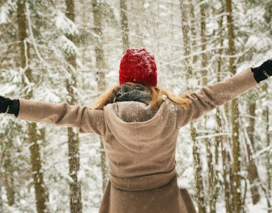 Mujer con gorro rojo y abrigo de espaldas, con los brazos abiertos en un bosque nevado